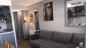 Ein modernes Wohnzimmer mit grauem Sofa, einem Essbereich mit Tisch und Stühlen, einer Küchenzeile mit Herd und einem großen Spiegel, der den Raum reflektiert.