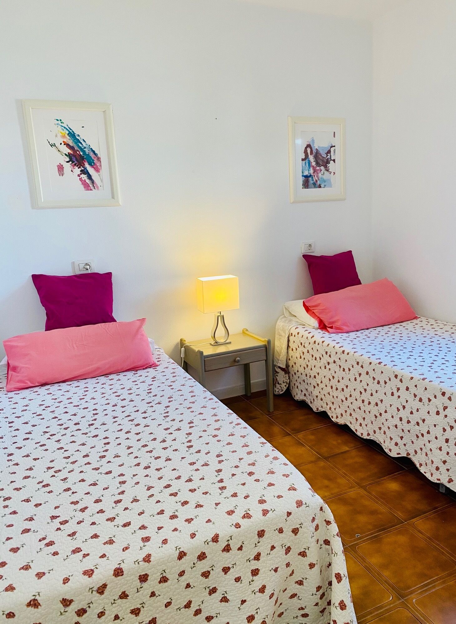 A room with two beds, each with a floral bedspread and pink pillows. There are two framed artworks on the walls, a nightstand with a lamp, and a small drawer.
