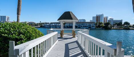 A wooden pier leading to a gazebo with a view of a body of water and buildings in the background.