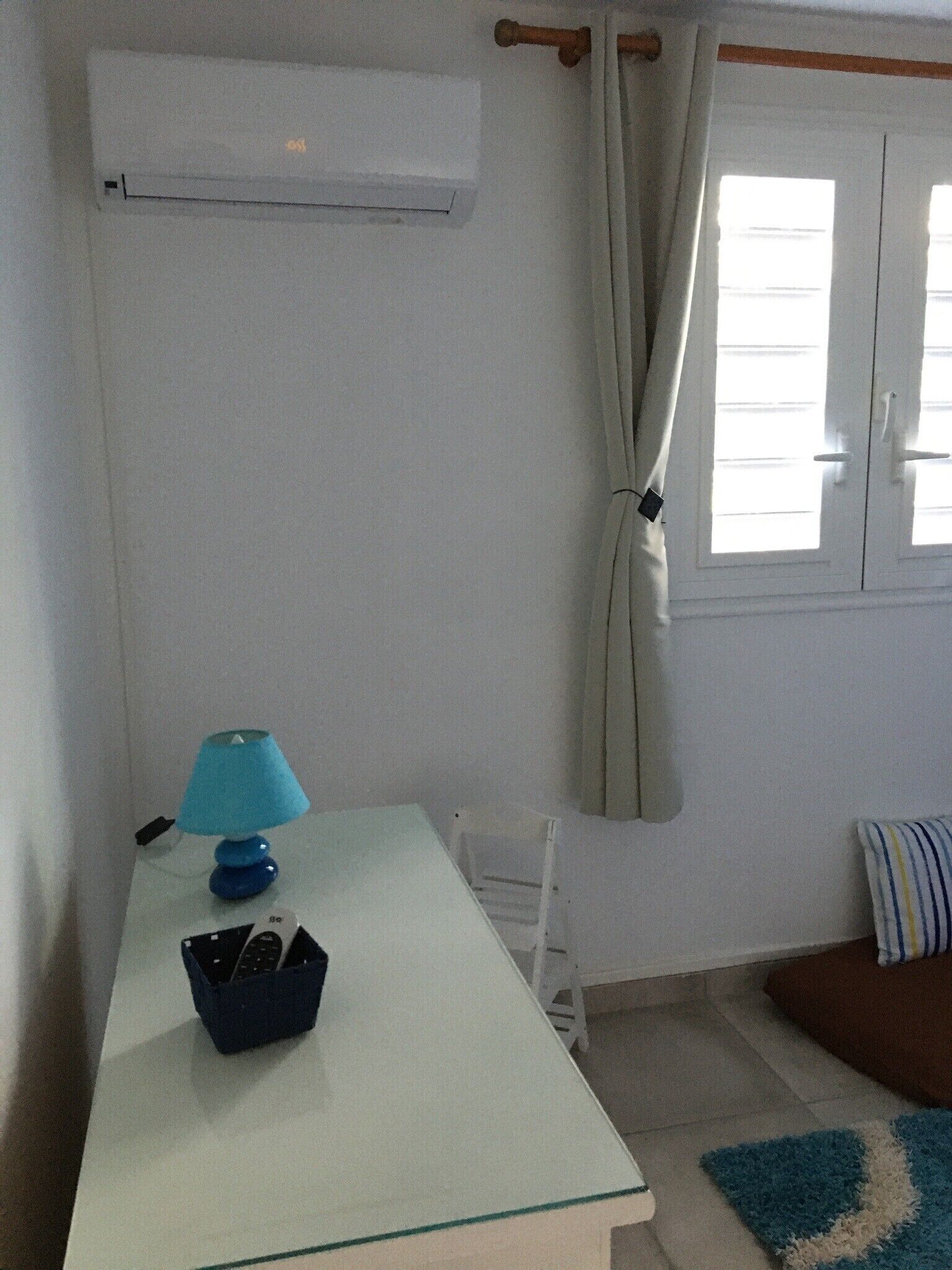 Una habitación con una mesa blanca, una lámpara azul, una silla blanca, un sofá marrón, un cojín a rayas y una ventana con cortinas.