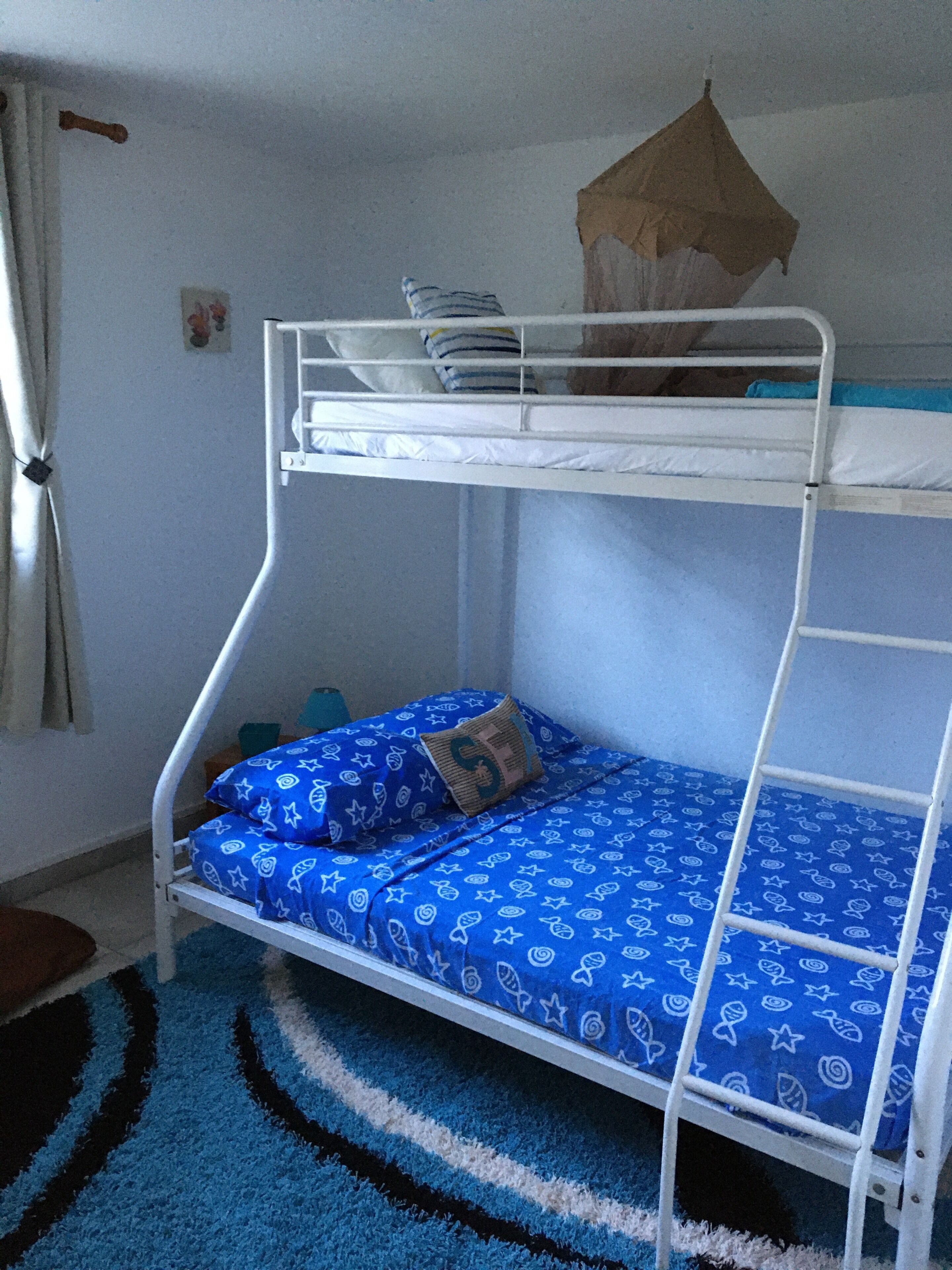 Uma cama beliche com edredom e almofadas estampadas em azul, estrutura metálica branca e tapete listrado em azul e branco no chão.