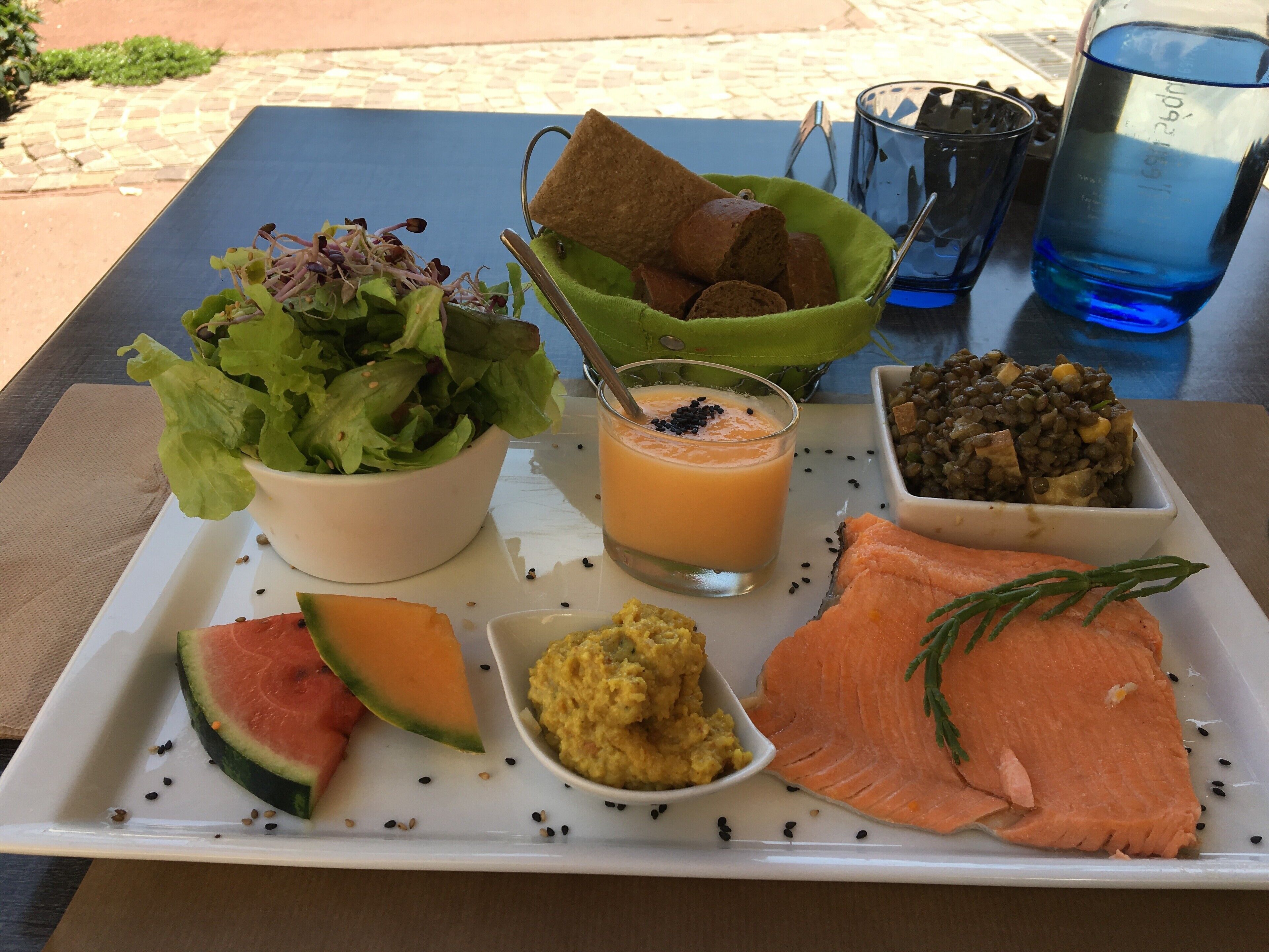 A meal tray with salmon, watermelon, a bowl of salad, a glass of smoothie, and a bowl of lentils.