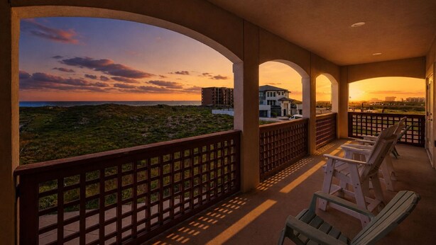 A balcony with a view of a sunset, a building in the background, and a fence. - Casa de la Perla, Ocean Views & Beach Boardwalk (Port Aransas)