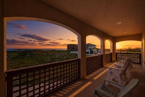 A balcony with a view of a sunset, a building in the background, and a fence. - Casa de la Perla ☀️ Boat Parking, Ocean View, Elevator & Beach Boardwalk | Pet Friendly (Port Aransas)