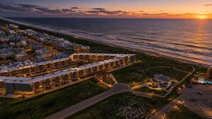 A coastal hotel with multiple buildings, a parking area, and a view of the ocean at sunset. - Casa de la Perla, Ocean Views & Beach Boardwalk (Port Aransas)
