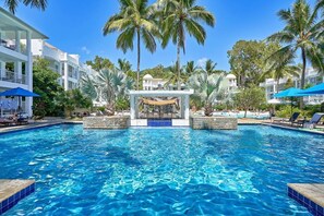 A swimming pool with a central water feature, surrounded by palm trees and lounge chairs.