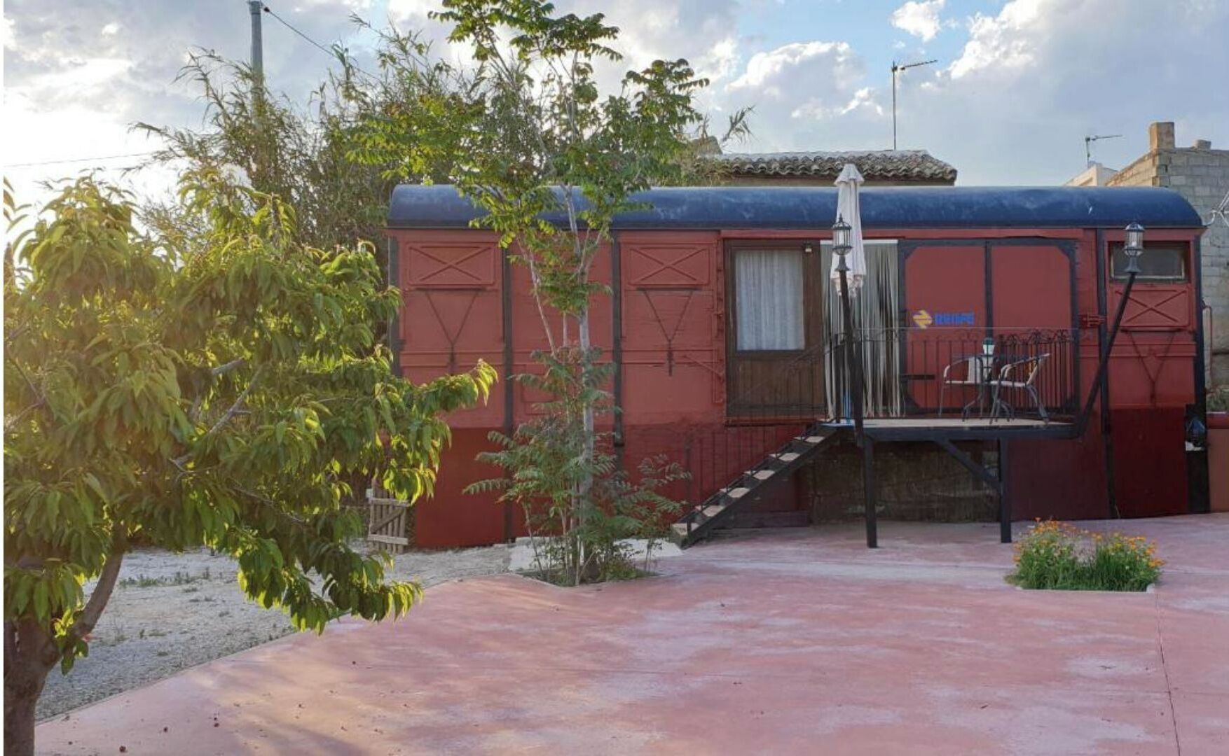 Um edifício vermelho com uma escadaria que leva a uma varanda. Há uma pequena área de jardim com plantas e uma árvore.
