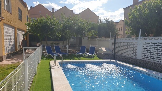 Ein Swimmingpool mit klarem, blauem Wasser, umgeben von einem weißen Zaun und einer Rasenfläche mit blauen Liegestühlen.