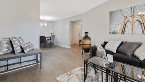 A modern living room with a sofa, a glass coffee table, and a dining area in the background. - The Gray Retreat - Luxury Condo (Fayetteville)