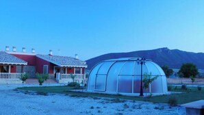 Una estructura de hivernacle amb teulada transparent, un edifici vermell amb porxo blanc al fons i un paisatge de muntanyes.