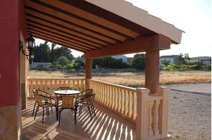 Un espacio de estar al aire libre cubierto con una mesa, sillas, barandilla y vistas a un área ajardinada con árboles y edificios al fondo.