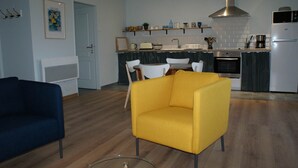 A modern living room with a sofa and a yellow armchair, a kitchen with a stove and oven, and a door leading to another room. - Gîte Blaen (CHERRUEIX)
