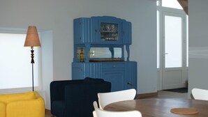 A room with a dining table, chairs, a blue cabinet with glass doors, a lamp, and a yellow sofa. - Gîte Blaen (CHERRUEIX)