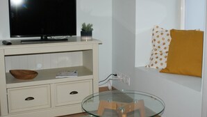 A modern living room with a flat-screen TV on a white cabinet, a glass coffee table, and a small wooden stool. - Gîte Blaen (CHERRUEIX)