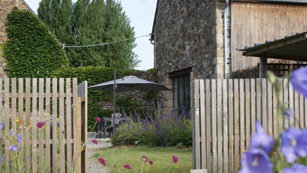 A wooden gate leading to a patio area with a table and chairs, surrounded by flowers and a stone building. - Gîte Blaen (CHERRUEIX)