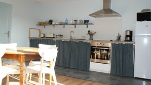 A modern kitchen with a wooden dining table, white chairs, a built-in oven, and a stainless steel range hood. - Gîte Blaen (CHERRUEIX)