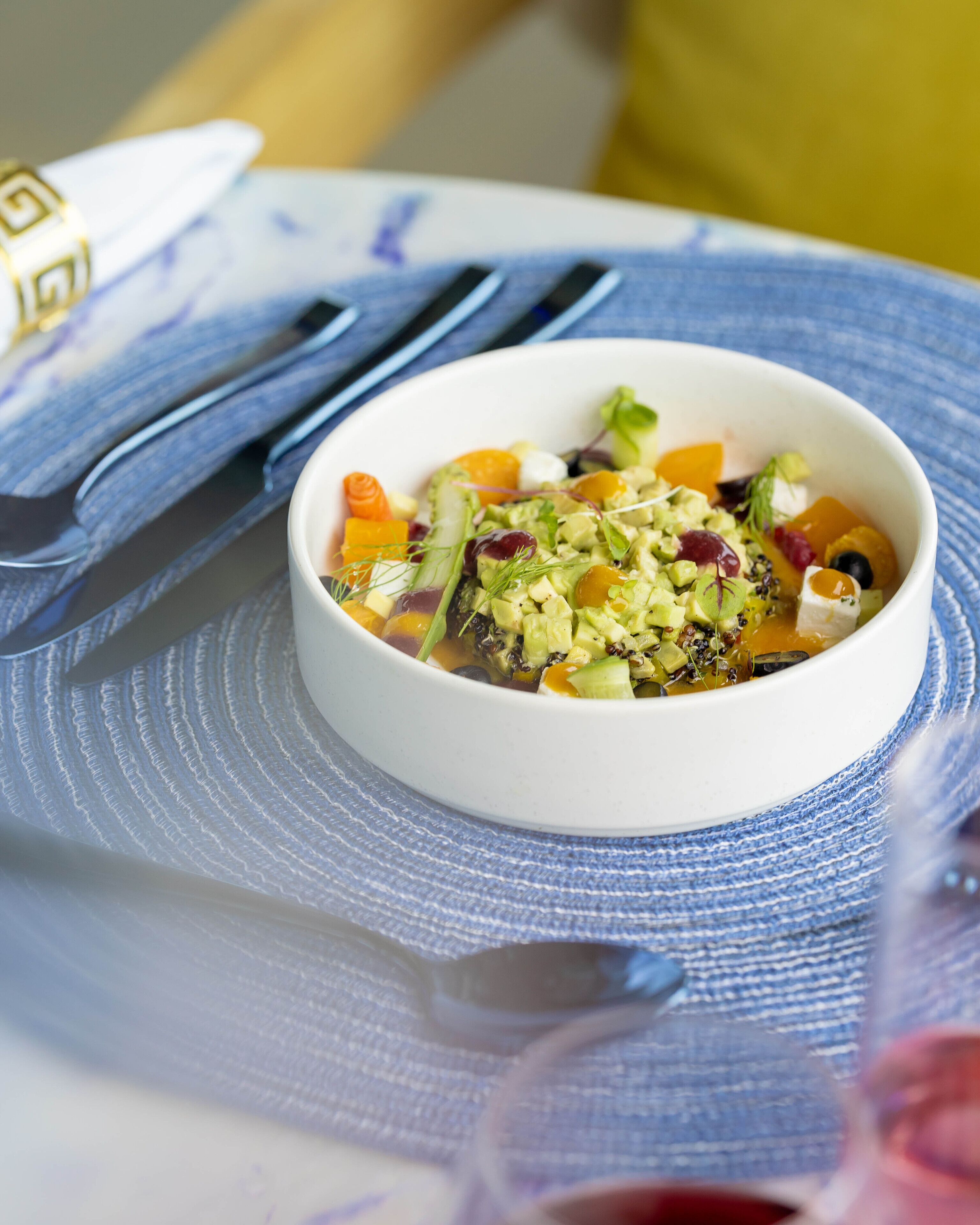 A bowl of salad on a blue placemat with a spoon and fork beside it.