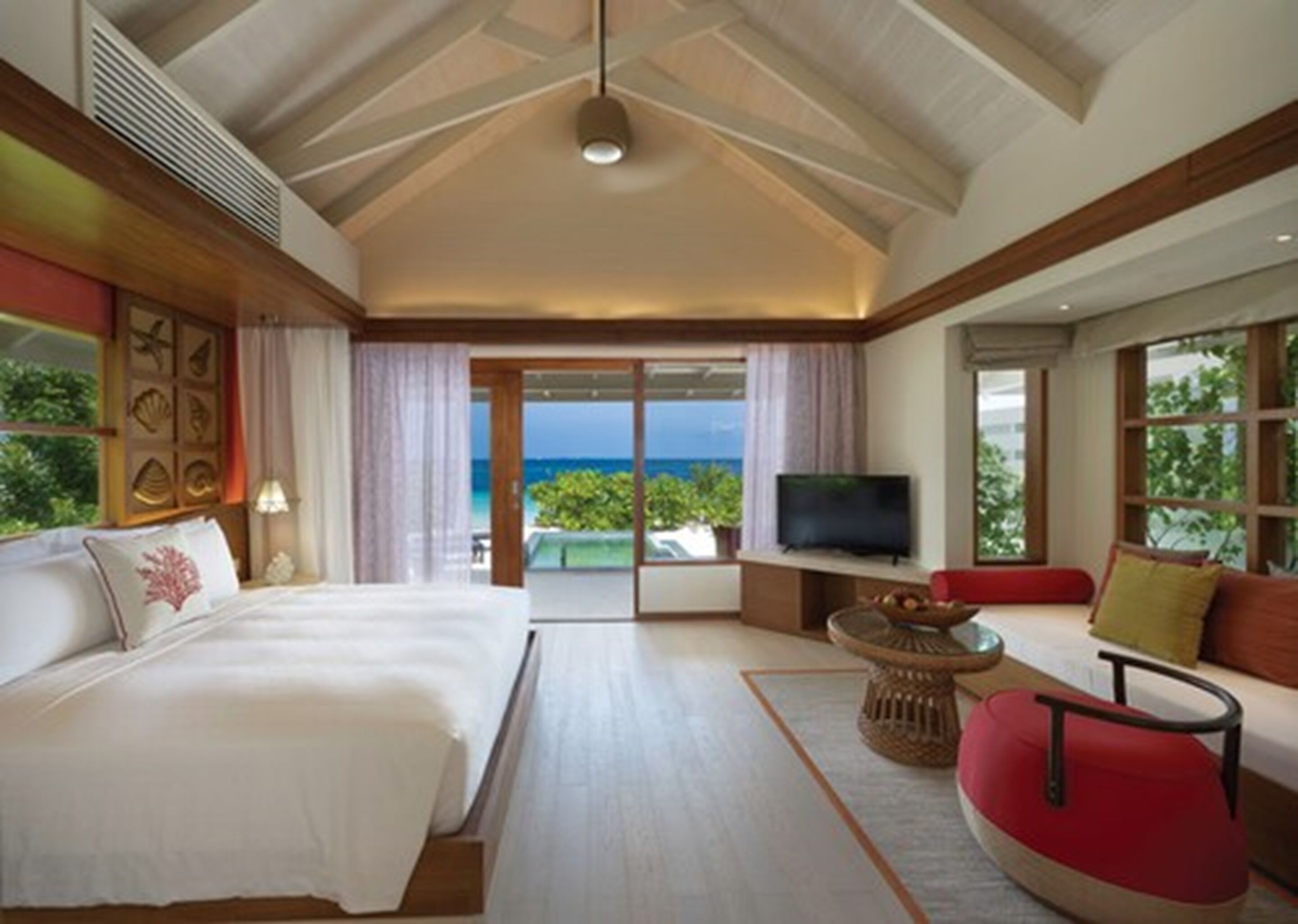 A spacious bedroom with a large bed, a TV, a seating area with a red chair, and a view of the ocean through large windows.