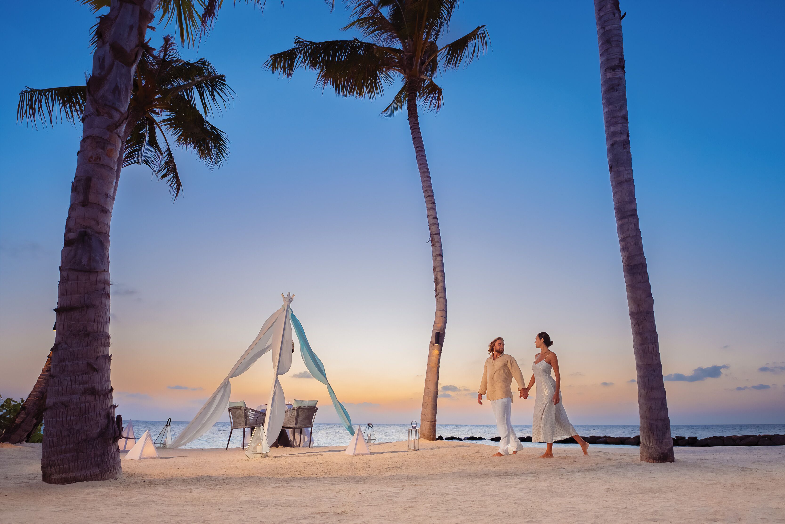 A beach scene with a tent, palm trees, and a couple walking.