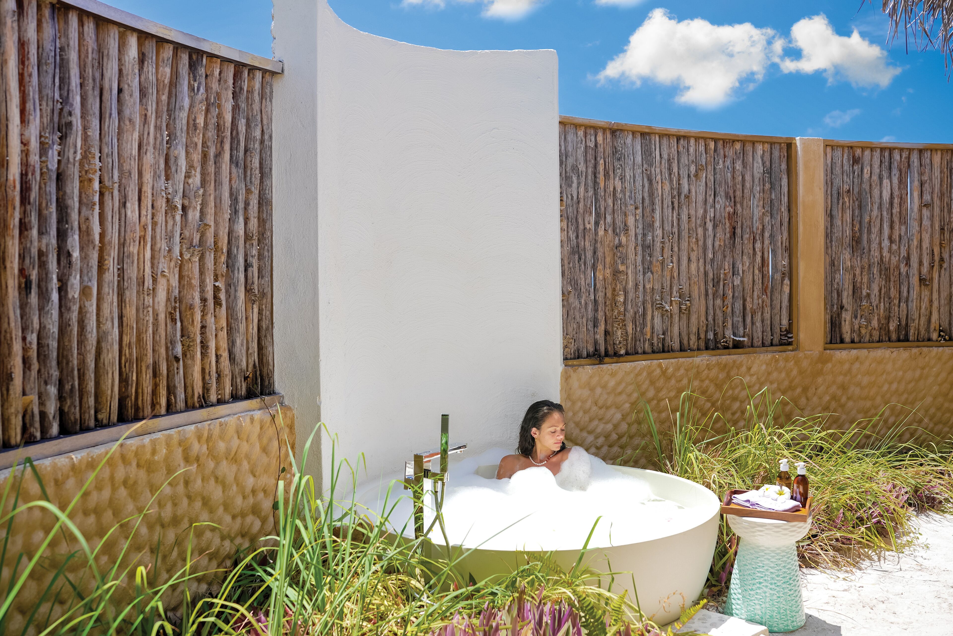 Eine Person entspannt in einer großen Freiluftbadewanne, umgeben von natürlicher Vegetation und Holzzäunen.