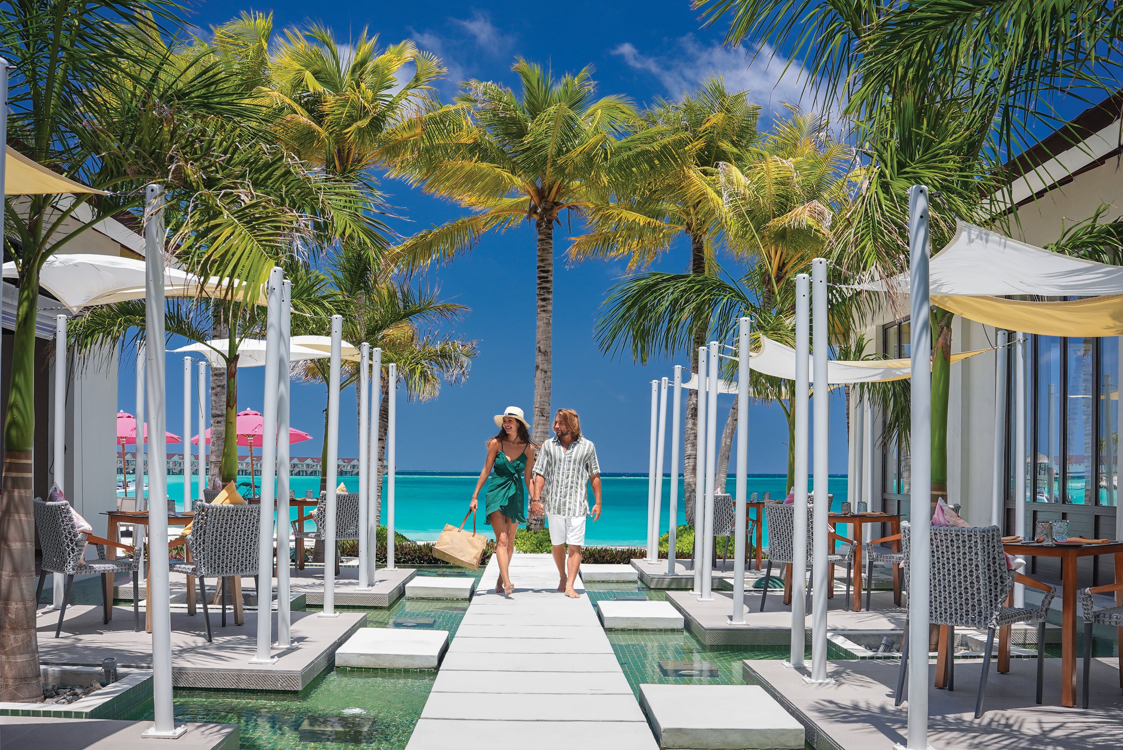 Una pareja caminando por un sendero junto a la piscina, con palmeras y sombrillas, con vista a un mar azul y despejado.