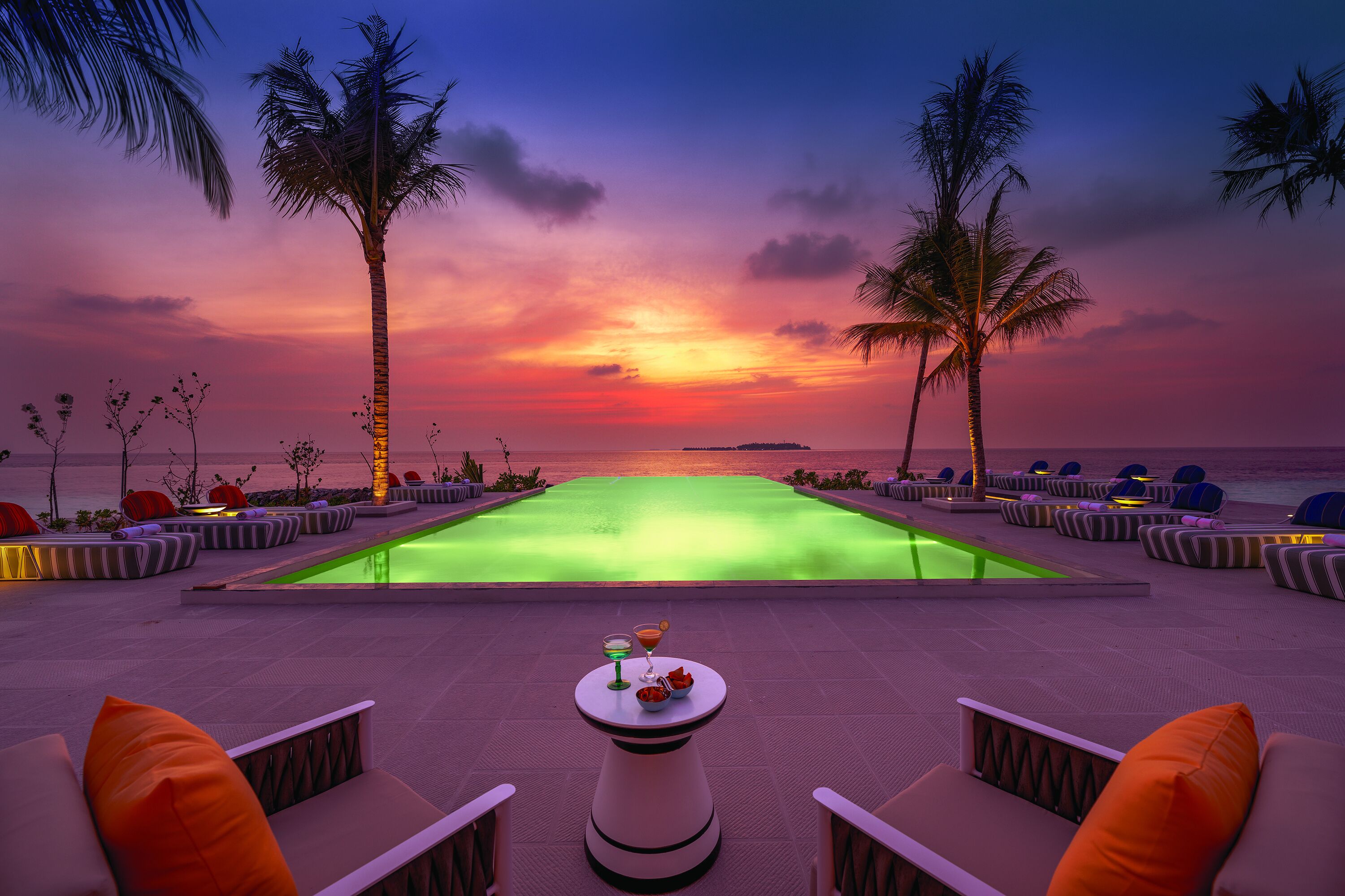 A poolside area with lounge chairs and a table with drinks, overlooking a sunset over the water.