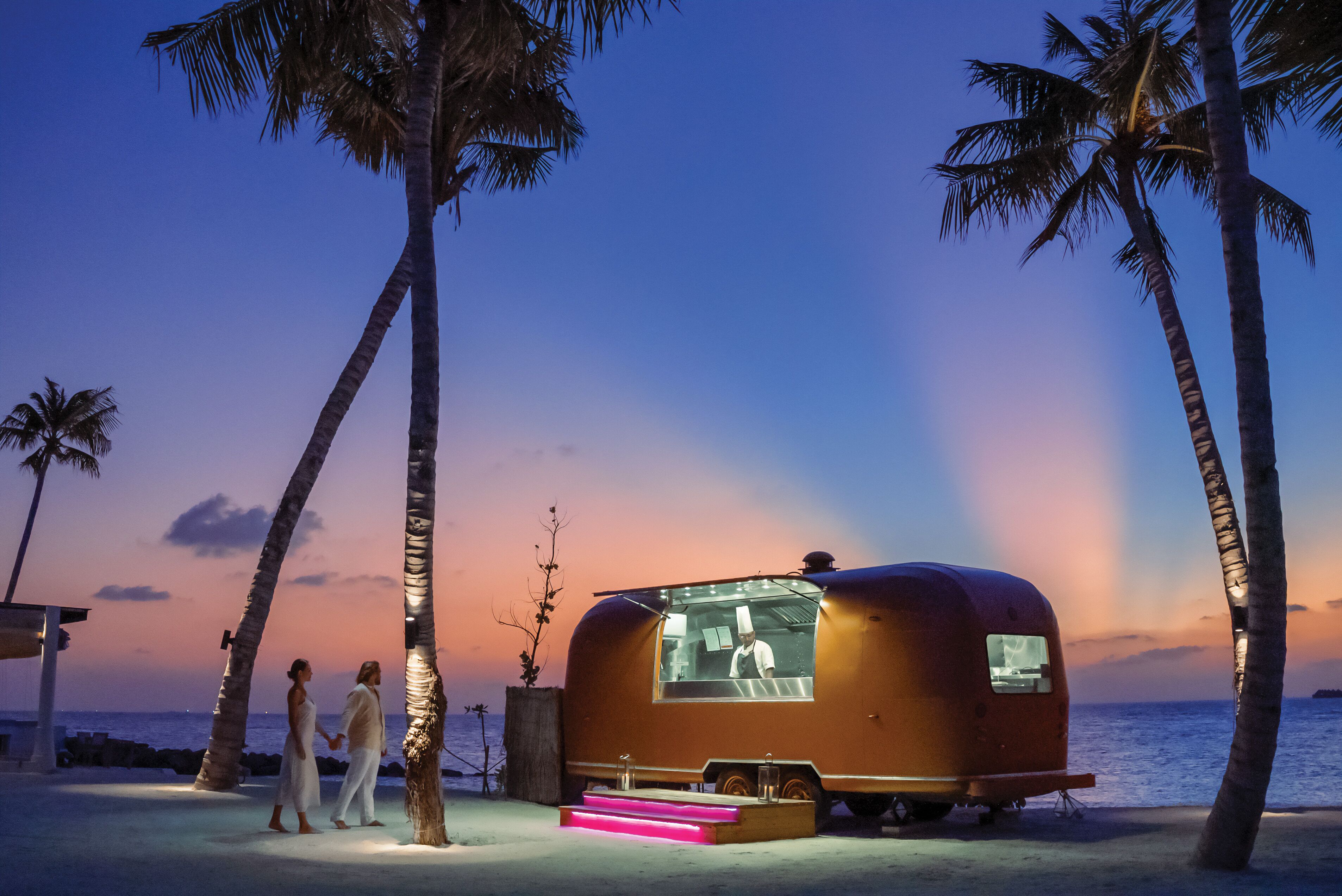 A vintage trailer with a chef visible through the window, parked on a beach with palm trees and a sunset in the background.