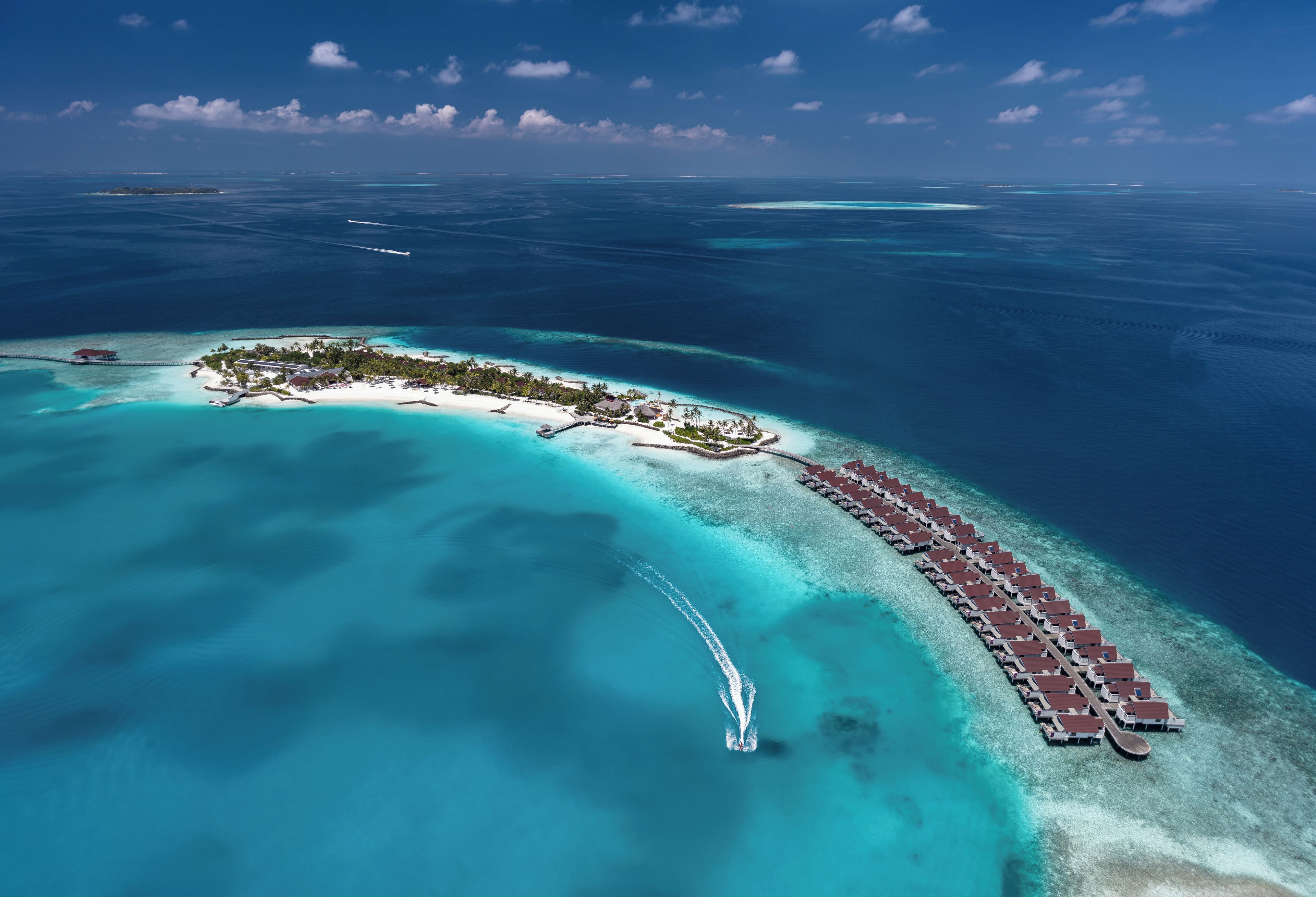 An aerial view of a resort island with overwater bungalows, clear turquoise waters, and a small mainland area with buildings.
