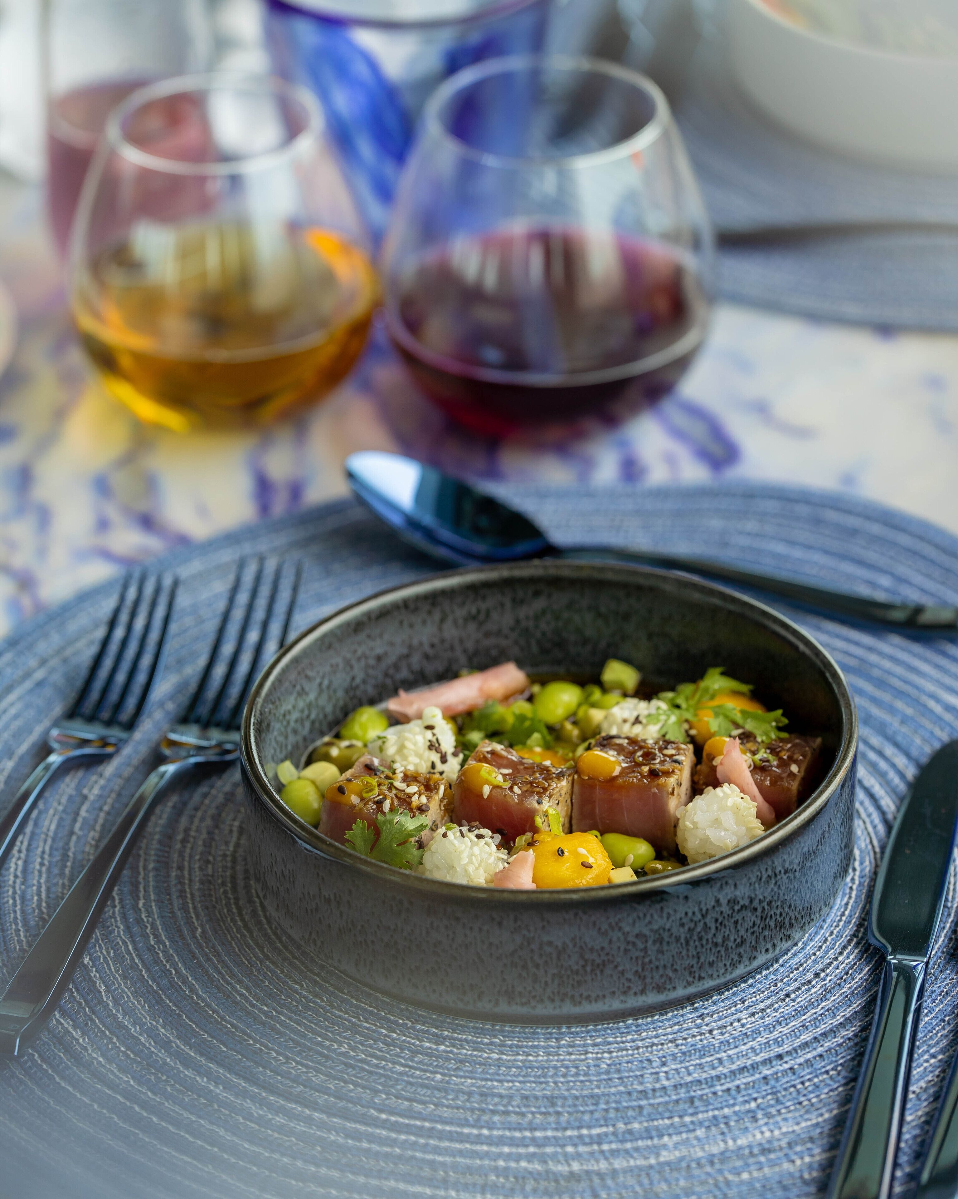 Un tazón de comida con arroz, verduras y carne, acompañado de cubiertos y vasos con bebidas sobre una mesa.