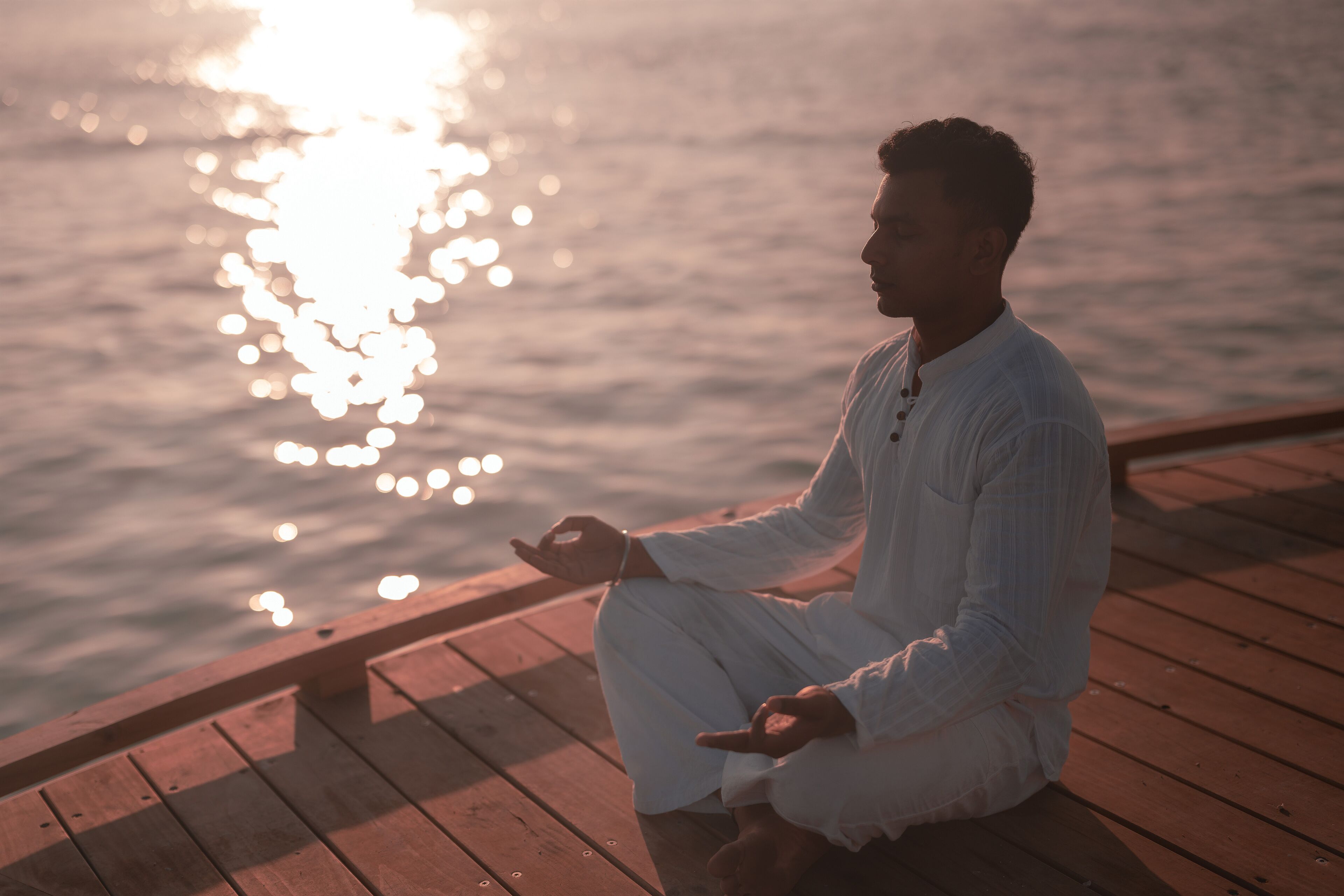 A person meditating on a wooden dock by a body of water at sunset.