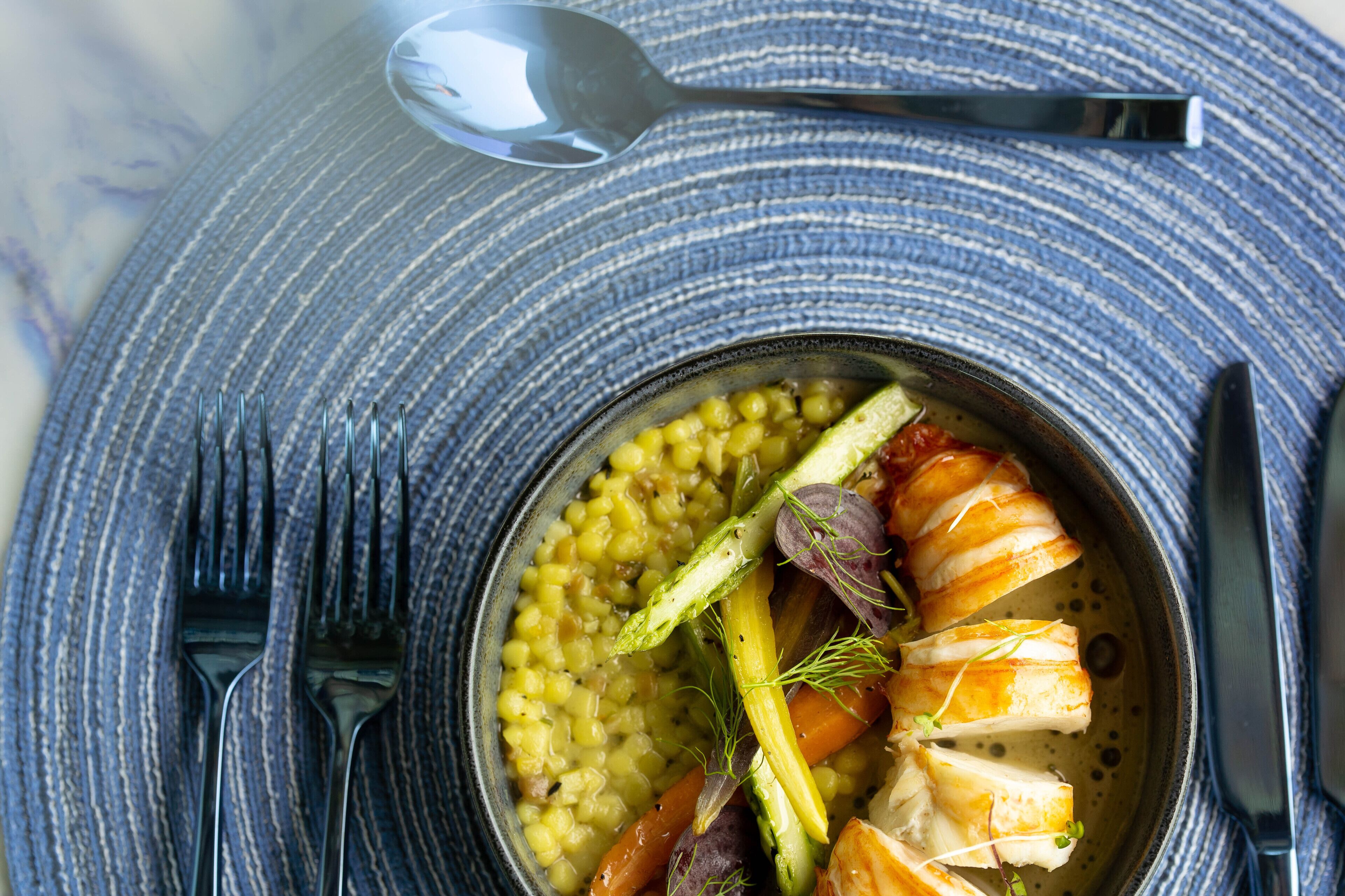 A bowl of food with corn, asparagus, and fish on a blue textured plate.