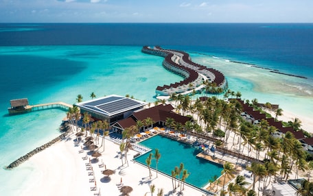 An aerial view of a resort with a swimming pool, beach, and overwater bungalows.