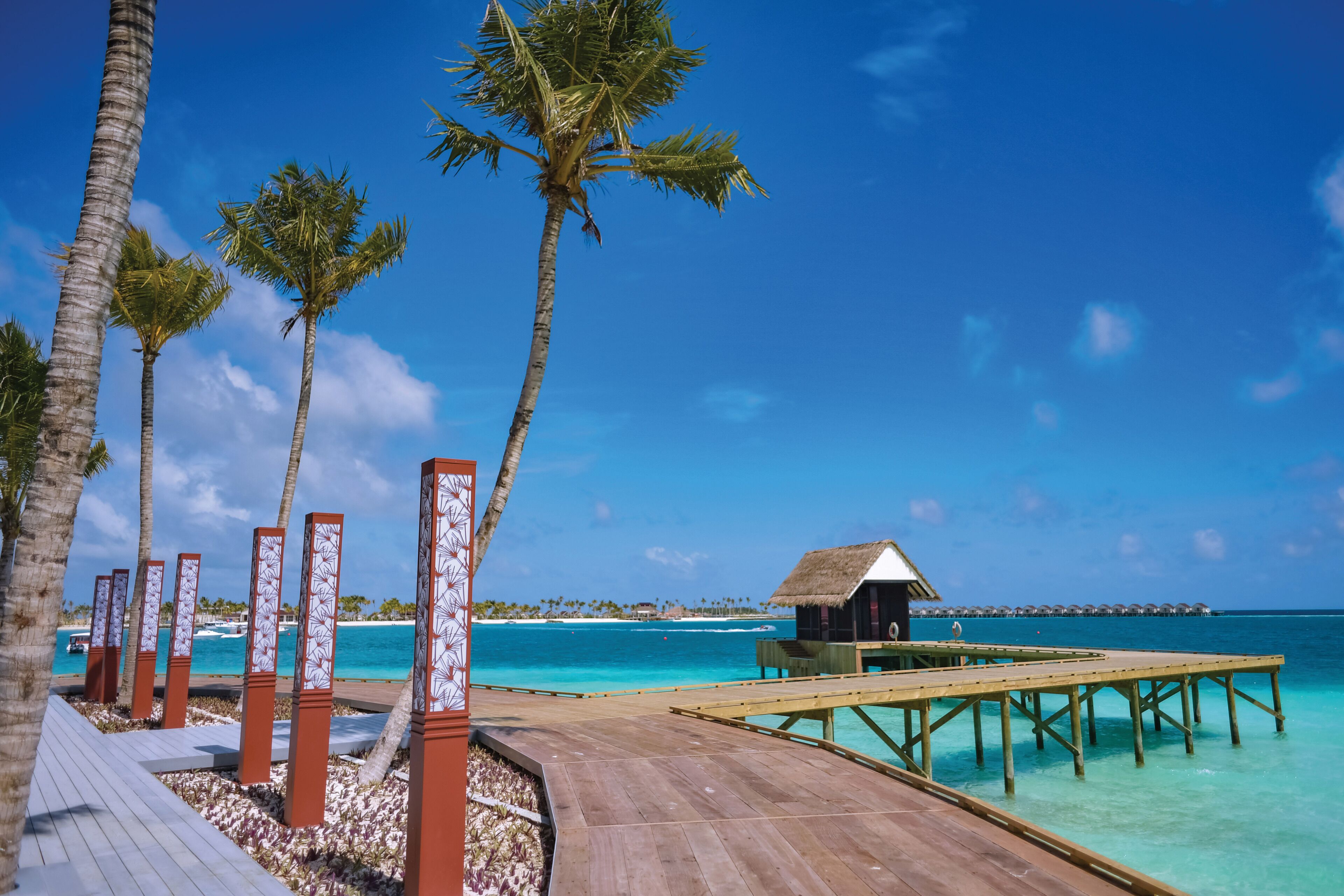 A wooden deck with palm trees, a thatched-roof structure, and a clear blue sea.