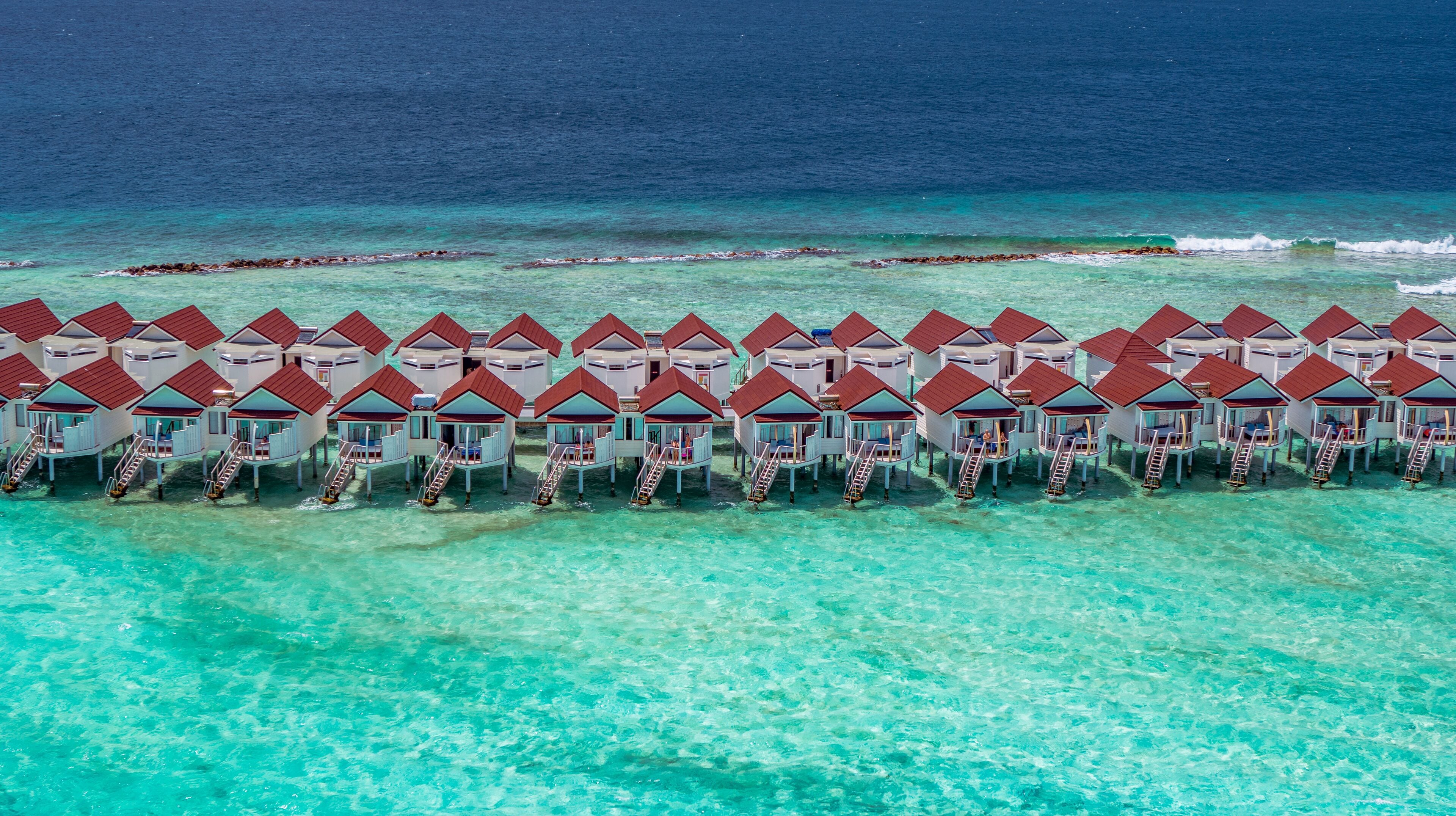 An aerial view of overwater bungalows with clear turquoise water and a distant coral reef.