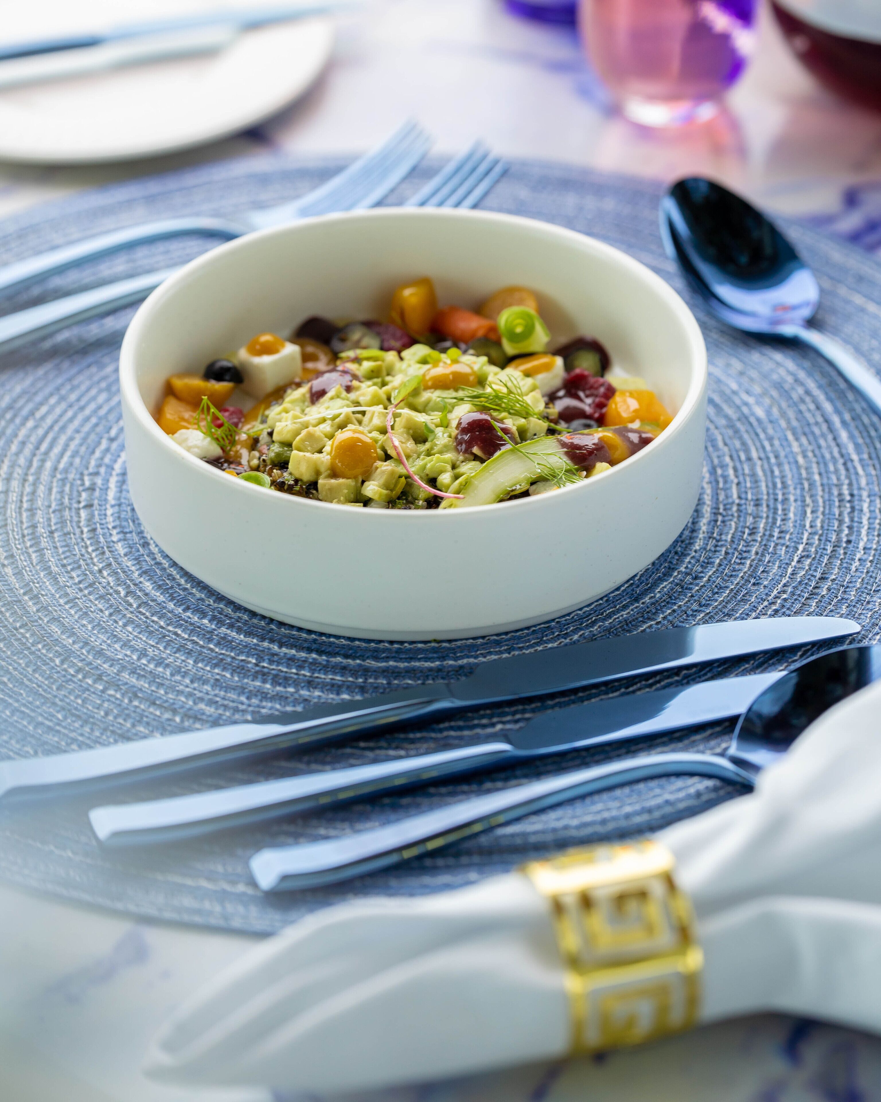 A bowl of salad on a table with a placemat, surrounded by cutlery and a napkin.
