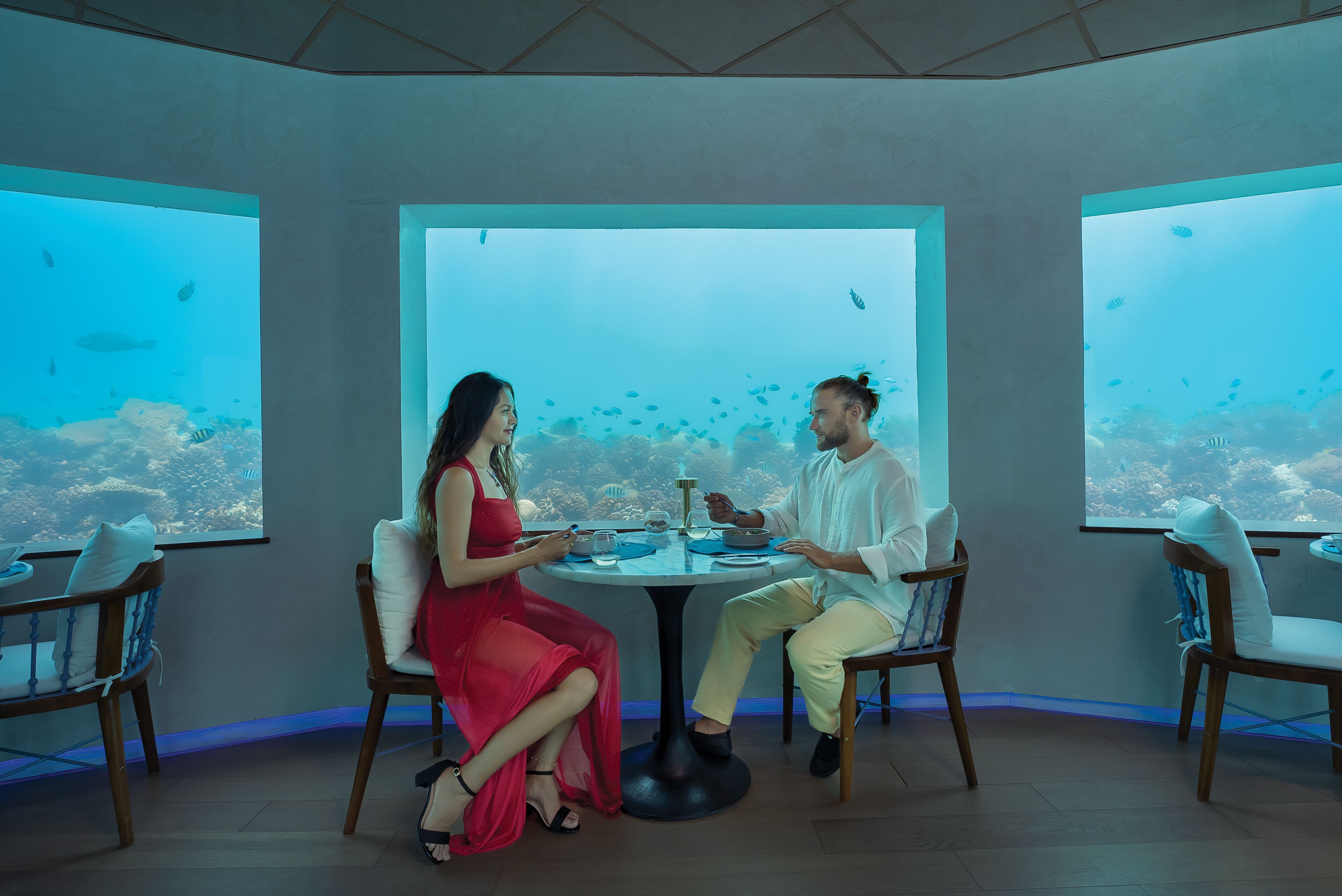 Un comedor con una mesa grande y sillas, ventanas amplias con vista al agua y un techo con diseño geométrico.
