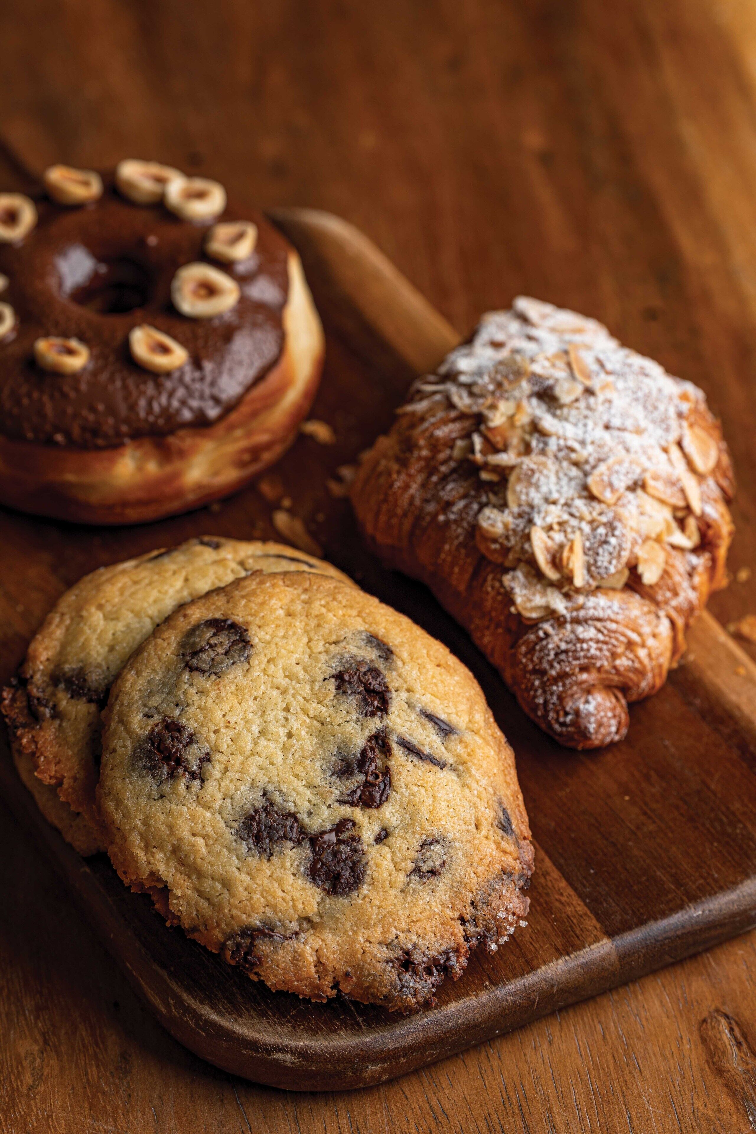 A wooden table with three baked goods: a chocolate-glazed donut, a chocolate chip cookie, and a powdered sugar-covered pastry.