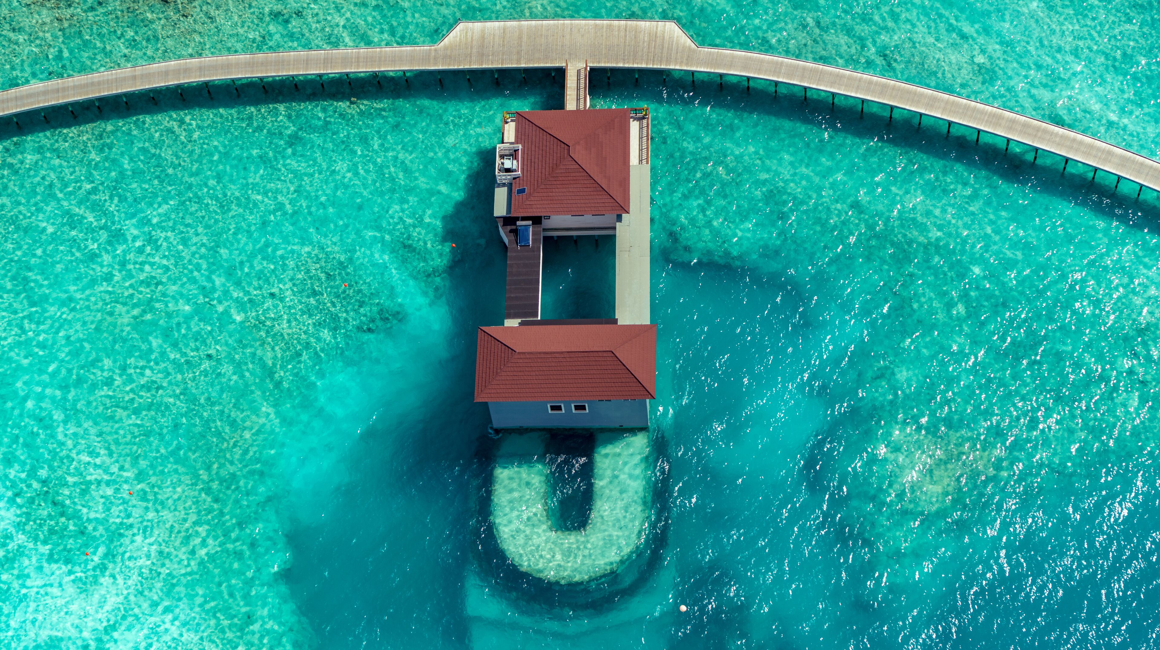 An aerial view of a resort with a long, narrow building with a red roof, connected to a pier extending into turquoise waters.