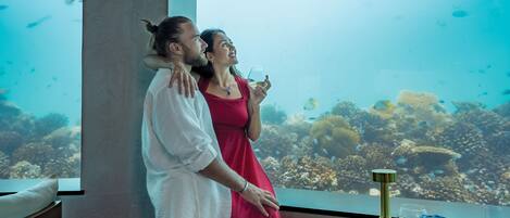A couple standing by a window with an underwater view, a dining table with plates and glasses, and a decorative lamp.