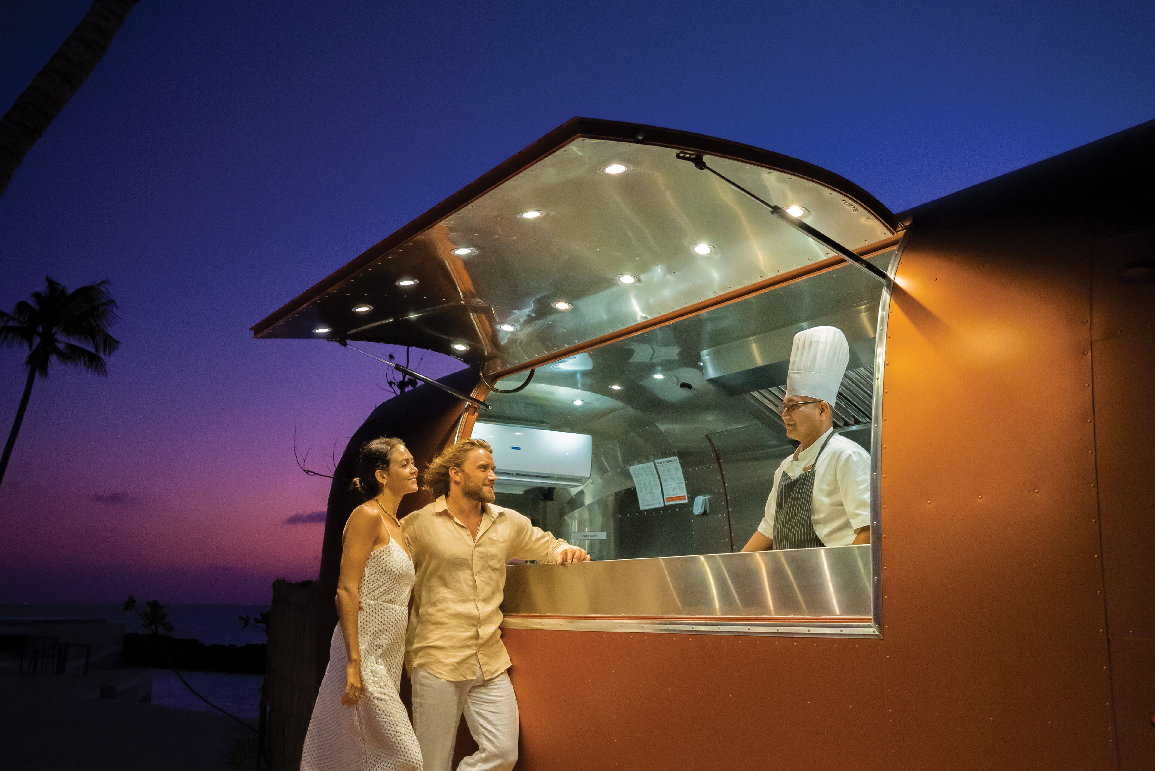 A food truck with a metal counter and a glass window, located near a beach with palm trees and a sunset in the background.