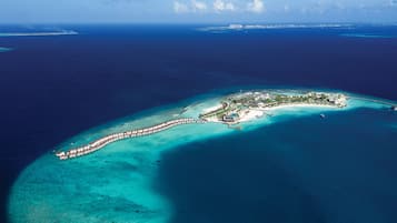 An aerial view of a tropical island with a long, white resort pier extending into turquoise waters, surrounded by clear blue sea and a small island with buildings and greenery.