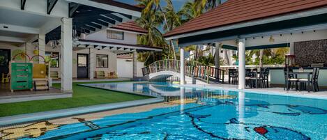 A swimming pool with a tropical design, surrounded by a covered area with seating and a view of palm trees.