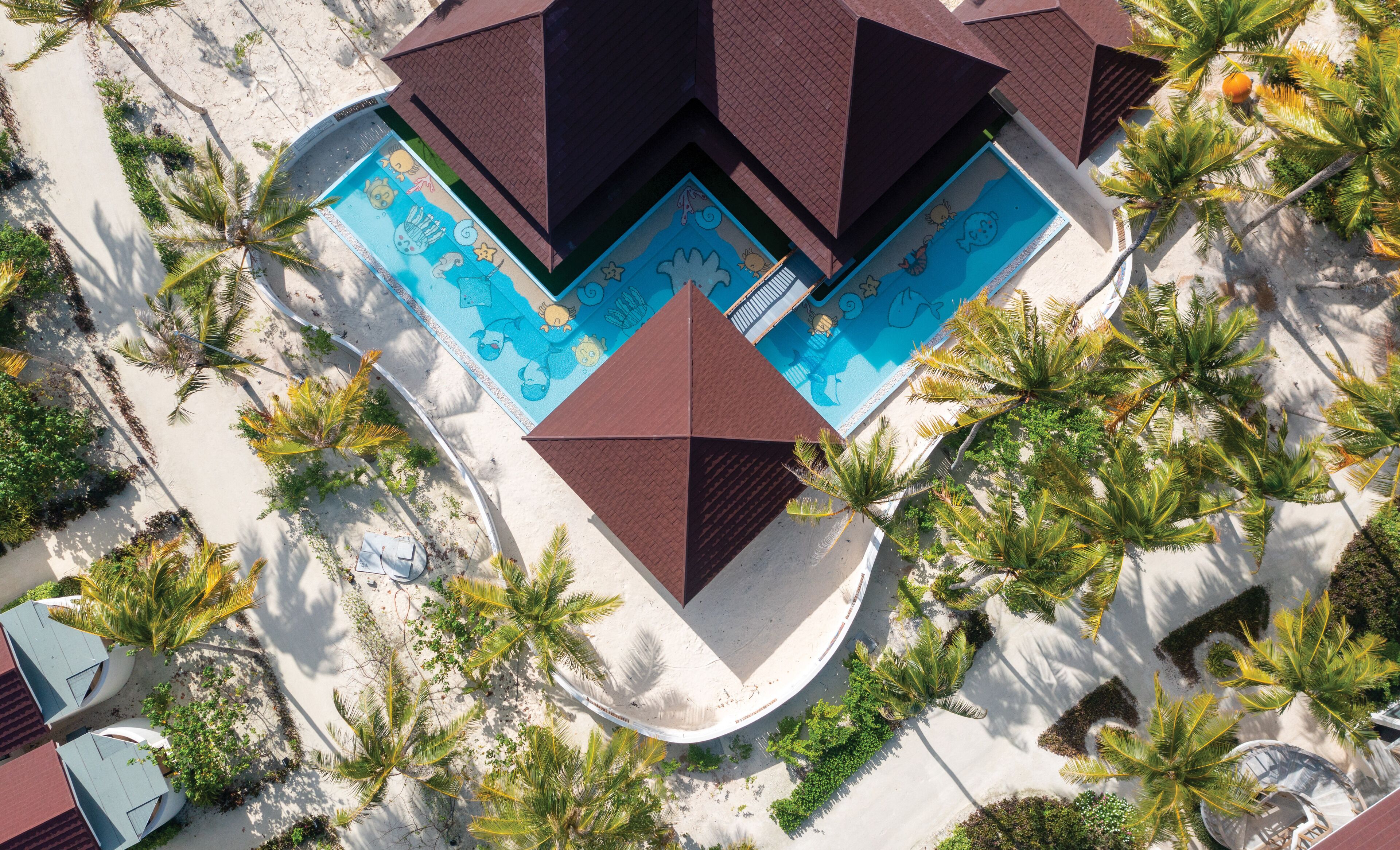 An aerial view of a resort with a central swimming pool surrounded by palm trees and a geometric pool deck design.