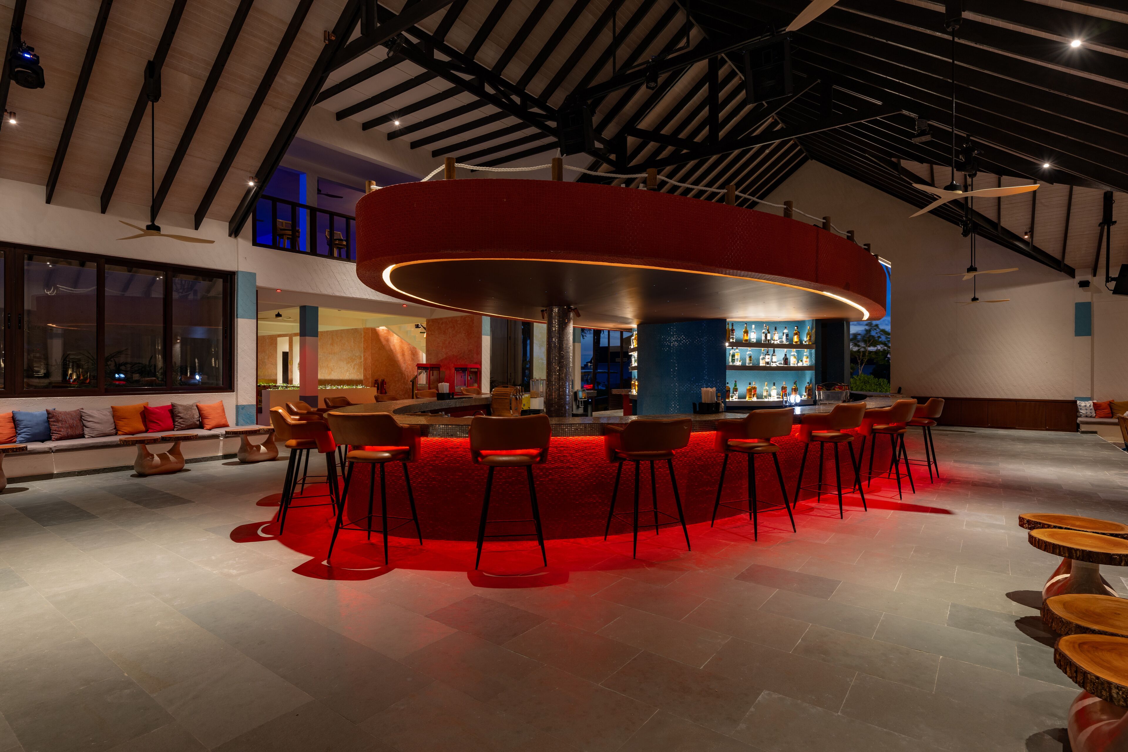 Un espace bar moderne doté d’un comptoir circulaire éclairé de lumières rouges, de nombreux espaces pour s’asseoir et d’un plafond haut avec des poutres apparentes.