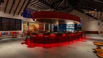 A modern bar area with a red-lit circular bar counter, multiple seating options, and a high ceiling with exposed beams.