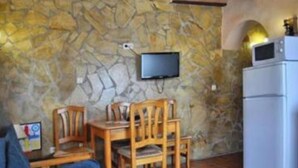 Une pièce chaleureuse avec un mur en pierre, un téléviseur à écran plat fixé au-dessus d’une table et de chaises en bois, et un réfrigérateur blanc.