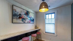 A modern kitchen with a marble countertop, pink bar stools, a large abstract painting, and a window with blinds. - Charming, modern, 2nd floor home-away-from-home (Augusta)