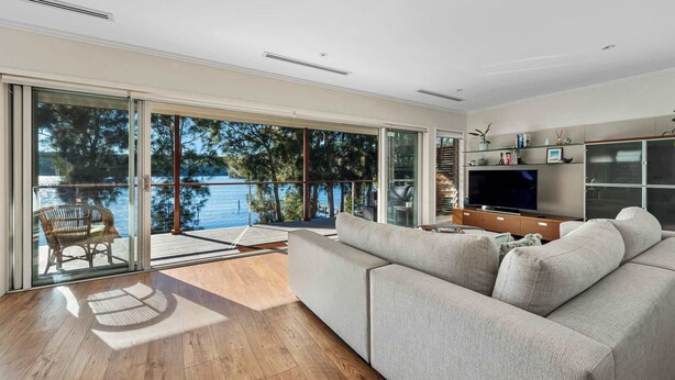 A modern living room with a large grey sofa, a flat-screen TV, and a glass door leading to a balcony with a view of a lake and trees.