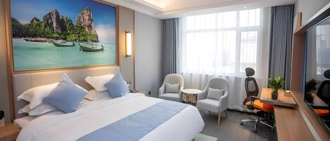 A hotel room with a large bed, a desk with a computer, a chair, a small table, and a painting of a coastal scene.