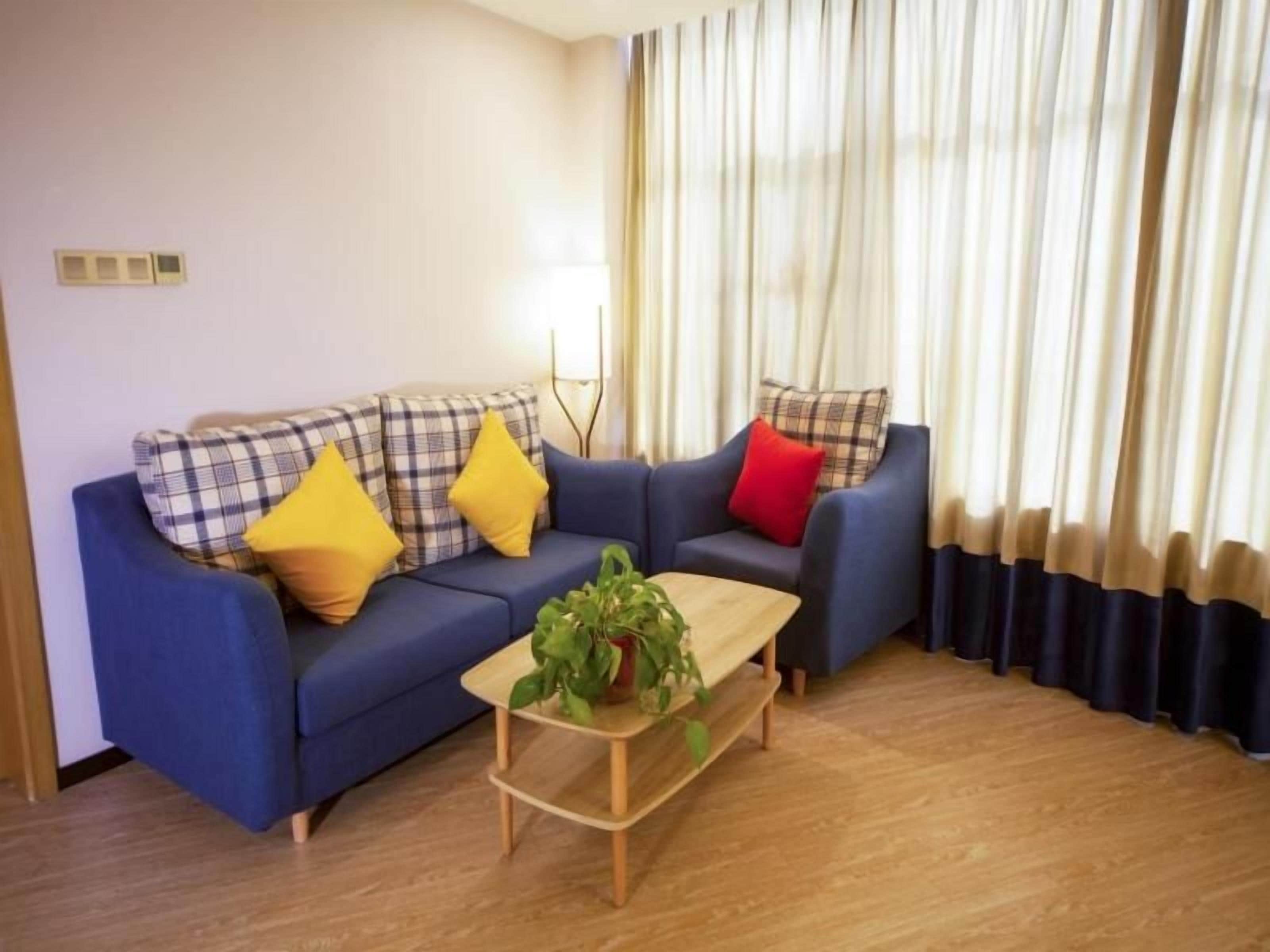 Una sala de estar con un sofá azul, cojines amarillos y rojos, una mesa de centro de madera con una planta, y vistas a una ventana con cortinas.